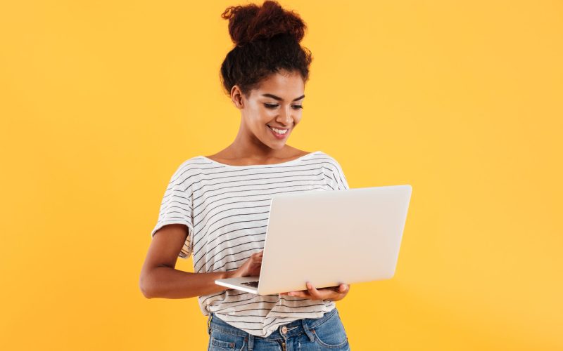 Young african positive cool lady with curly hair using laptop and smiling isolated over yellow